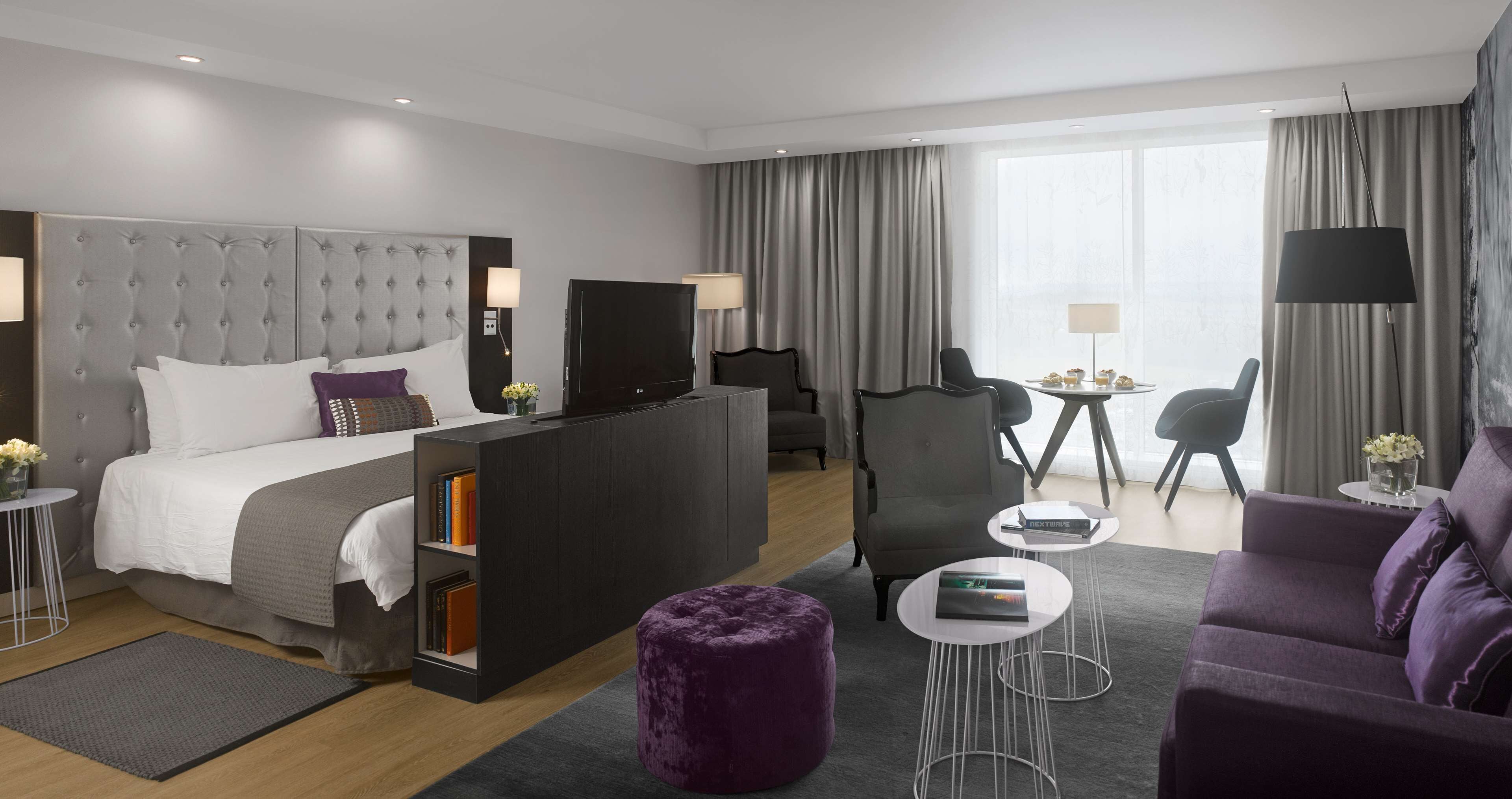 A modern hotel room with a large bed, a desk, two chairs, a sofa, and a TV.