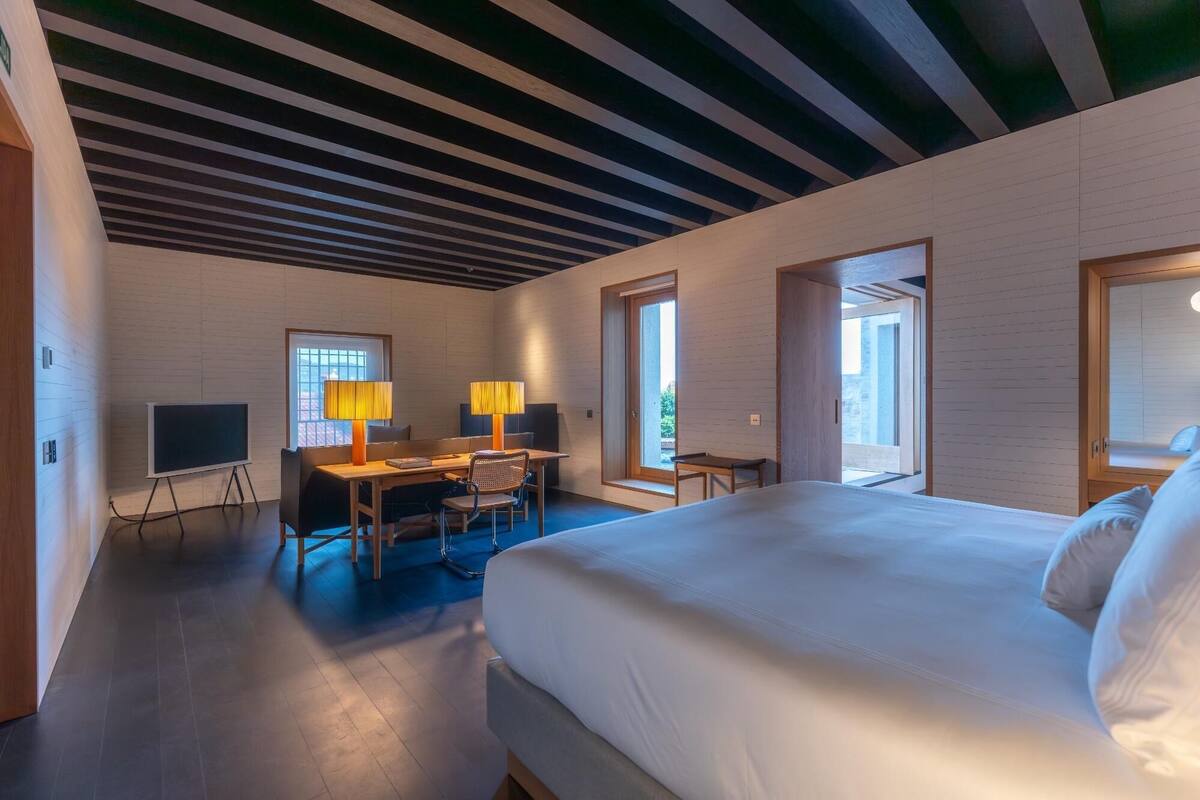 A hotel room with a bed, desk, and chairs, a TV, and large windows.
