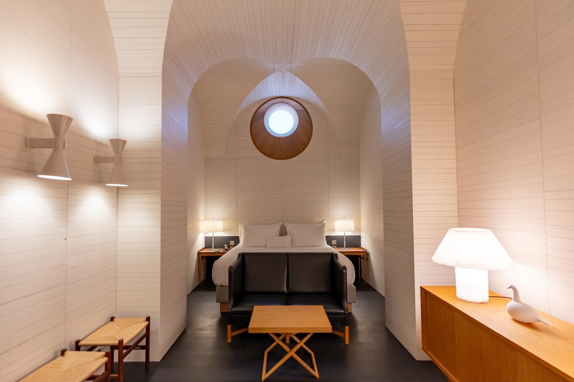 Una habitación de hotel moderna con una cama central, dos mesitas de noche, un tragaluz circular y un banco de madera.