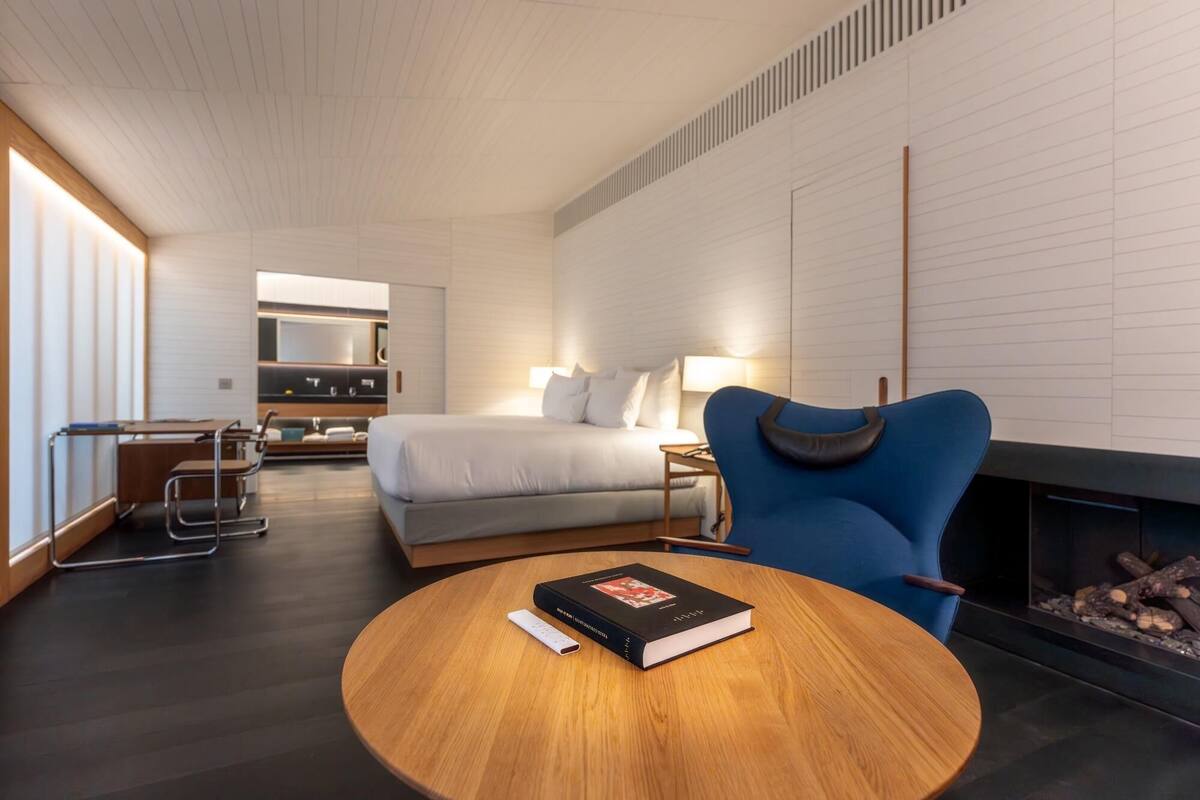 A modern hotel room with a bed, a desk, a chair, a fireplace, and a round wooden table.