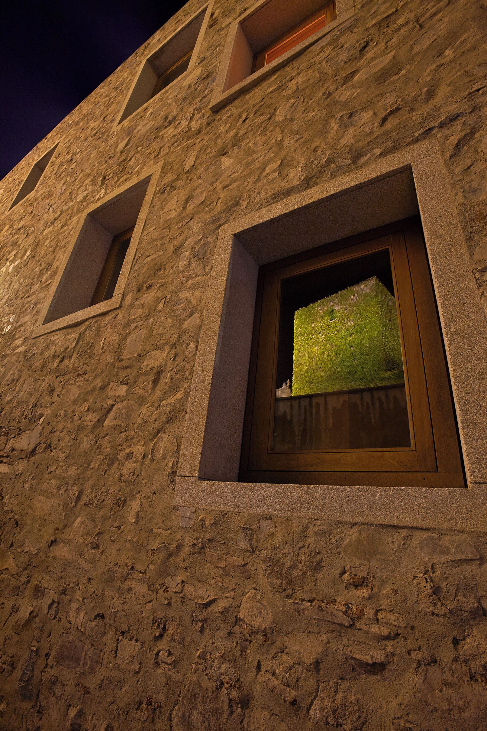 Fachada de edificio con revestimiento de piedra texturizada, una ventana con parte superior de tejas verdes y una ventana más pequeña a la izquierda.