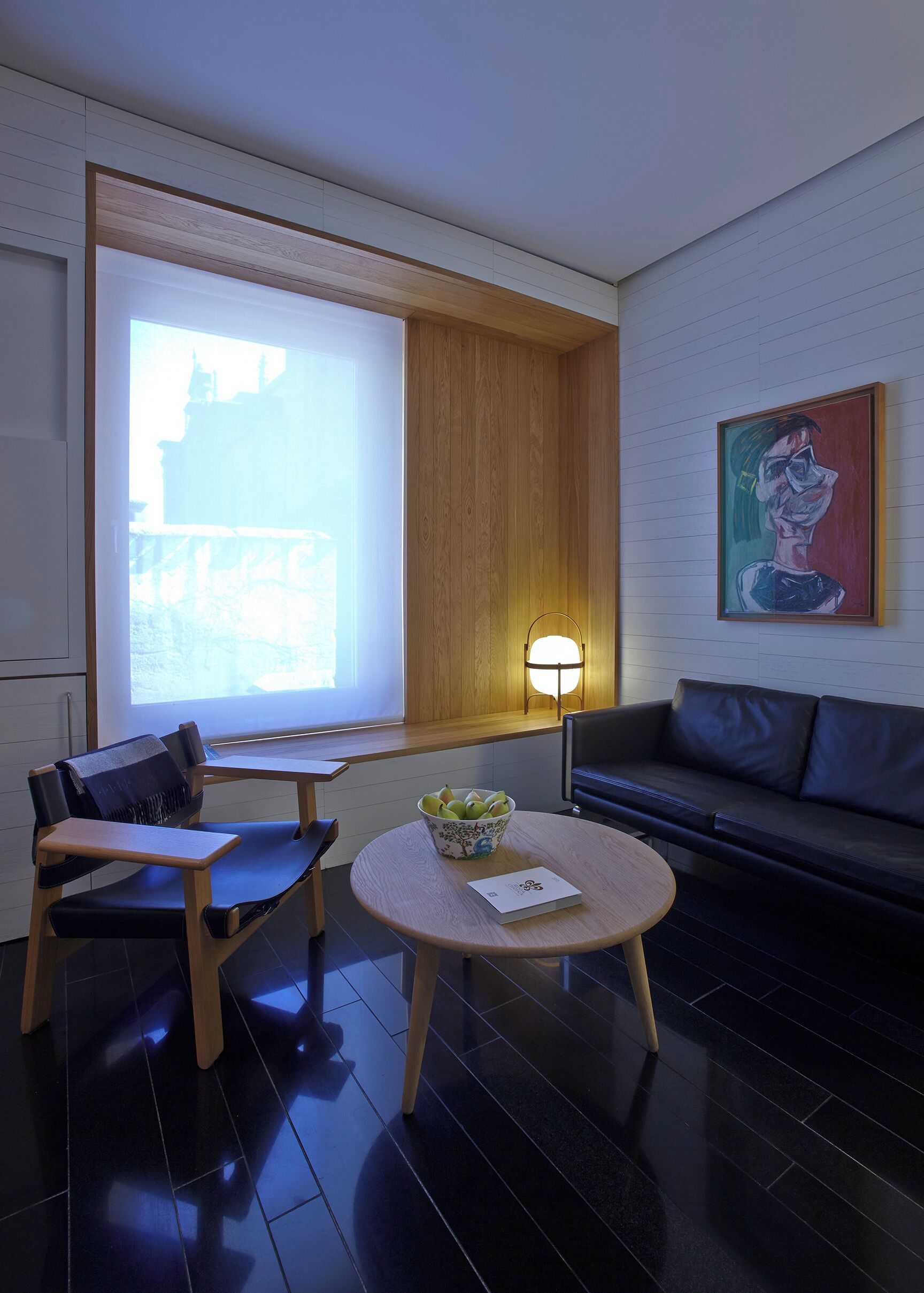 A modern room with a large window, a sofa, a table with a bowl of fruit, and a painting on the wall.