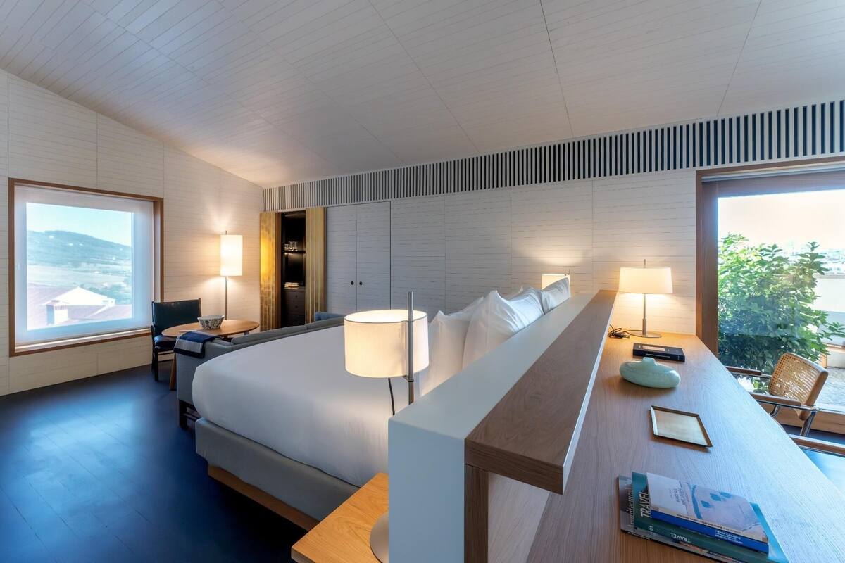 A modern hotel room with a large bed, bedside tables, a desk, and a view of the outdoors.