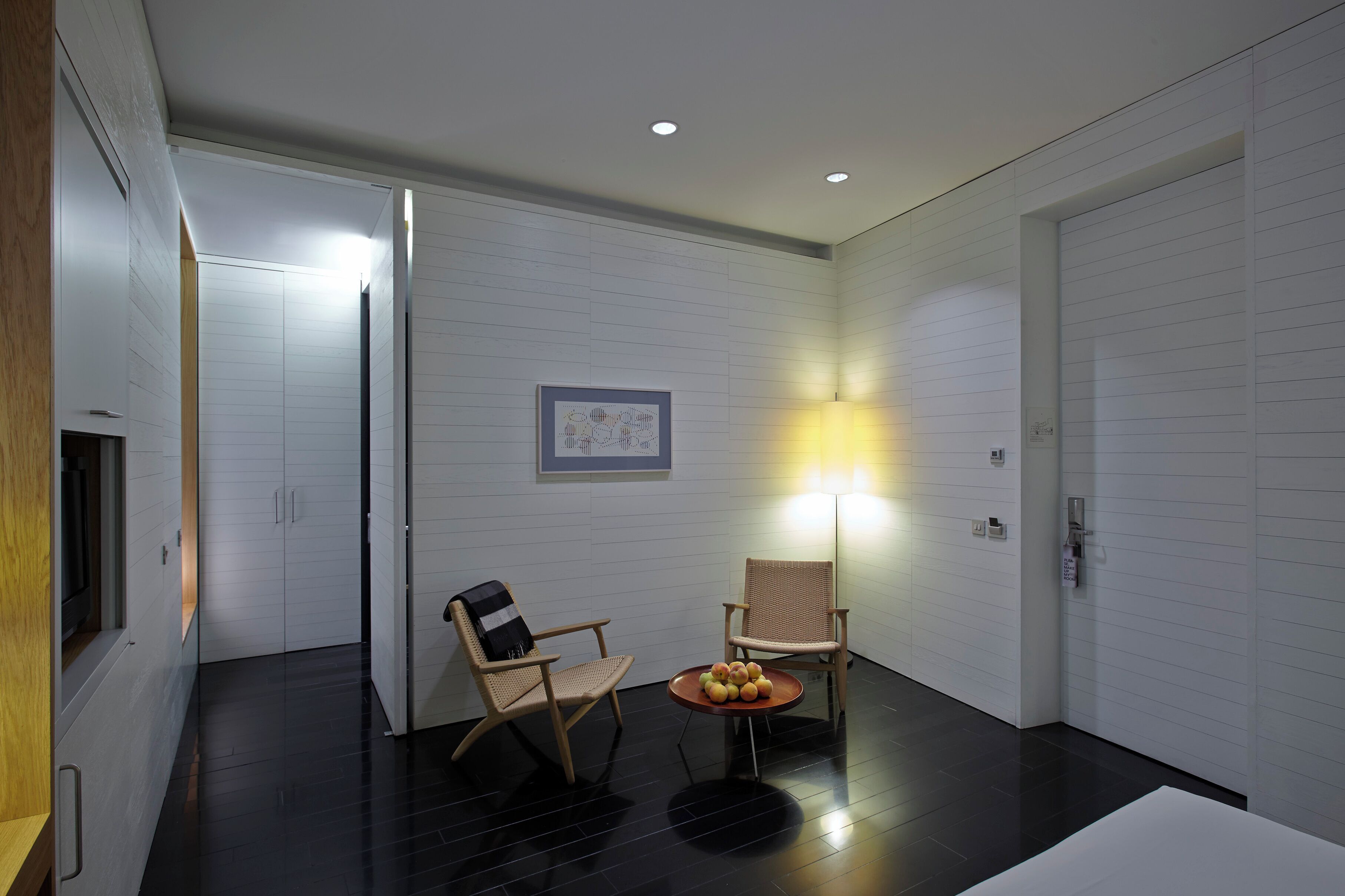Habitación moderna con paredes de paneles blancos, suelo de madera, dos sillones de mimbre, una mesita con fruta e iluminación empotrada en el techo.