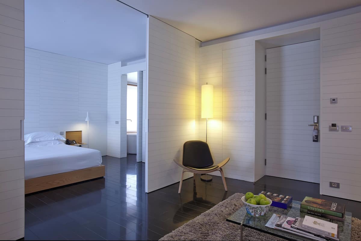 A modern hotel room with a bed, a chair, a lamp, a glass table with books and a bowl of fruit.