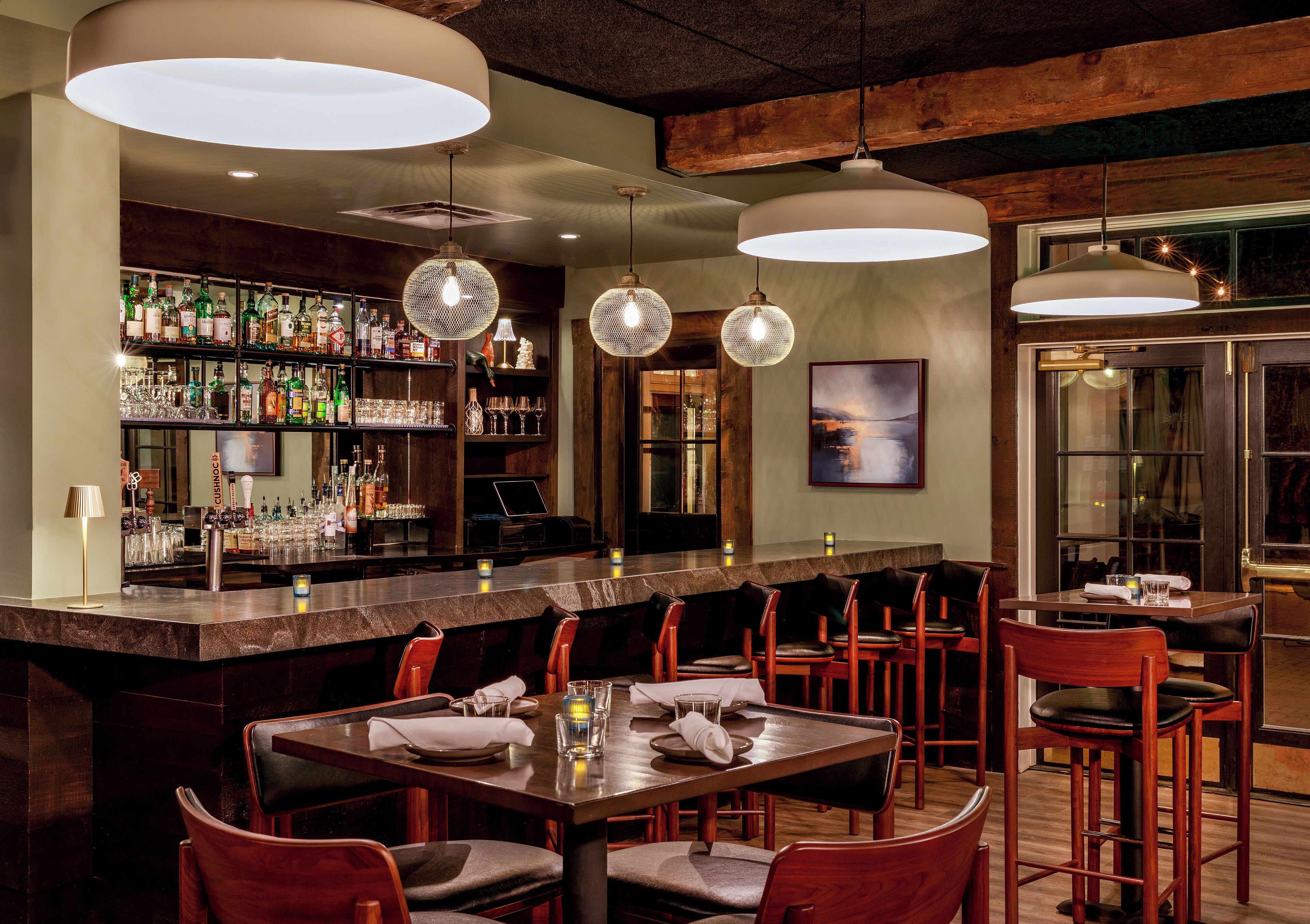 Un espace bar doté d’un long comptoir, de plusieurs luminaires suspendus et d’une variété de bouteilles et de verres disposés sur des étagères.