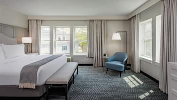 A hotel room with a large bed, a bench, a chair, a small table, and a lamp. There are windows with curtains and a view of the outside.