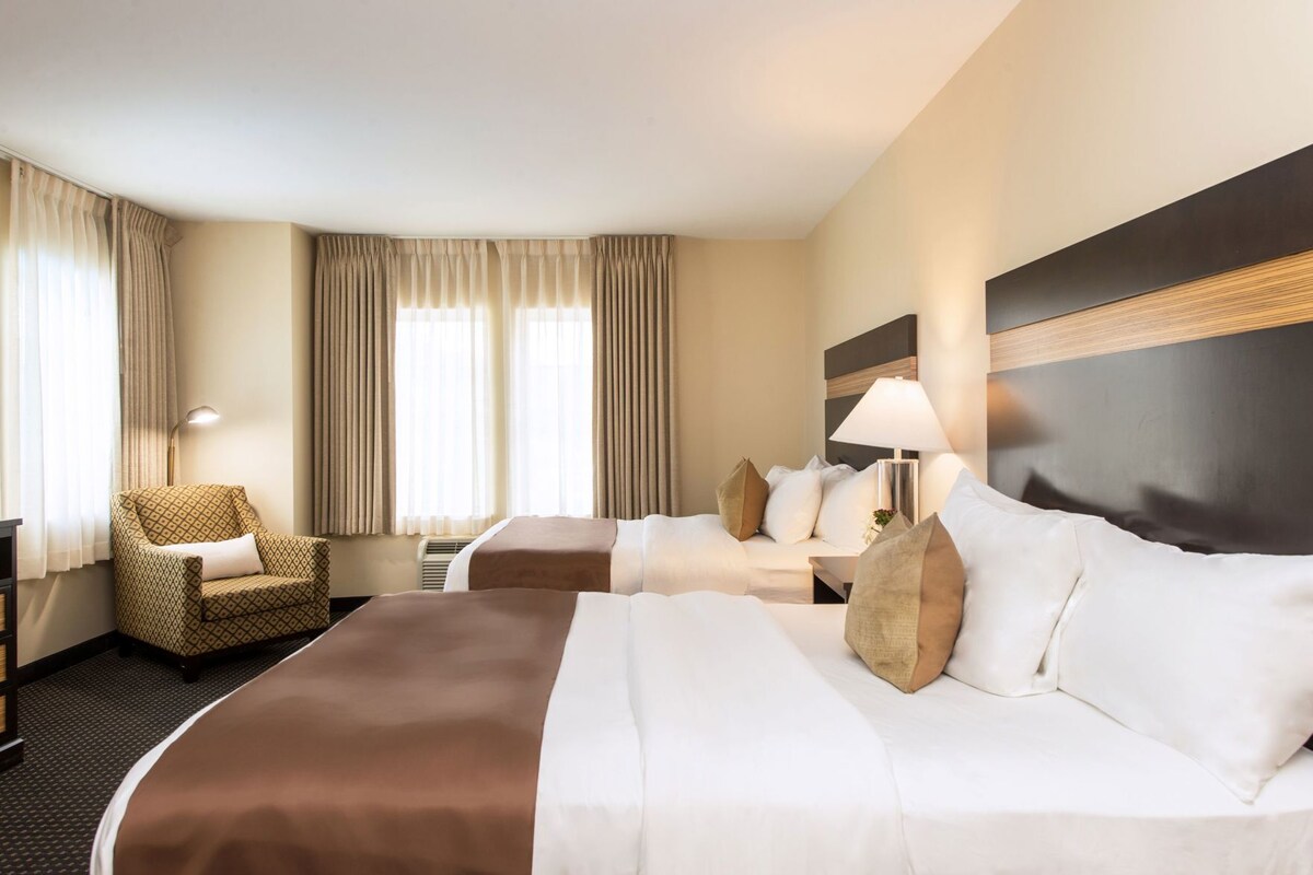 A hotel room with two beds, a chair, a desk, and a lamp.