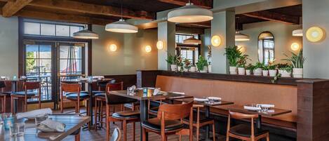 A restaurant interior with wooden tables and chairs, pendant lighting, and potted plants on the counter.