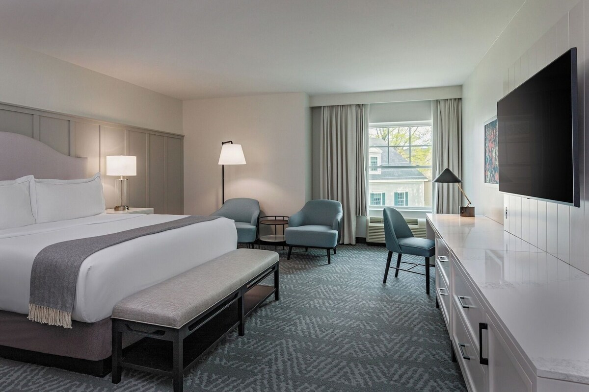 A hotel room with a bed, two armchairs, a small table, a bench, a TV, and a window with curtains.