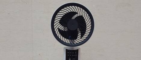 Un ventilador de pie con un panel de control, sobre una base blanca, frente a una pared de azulejos.