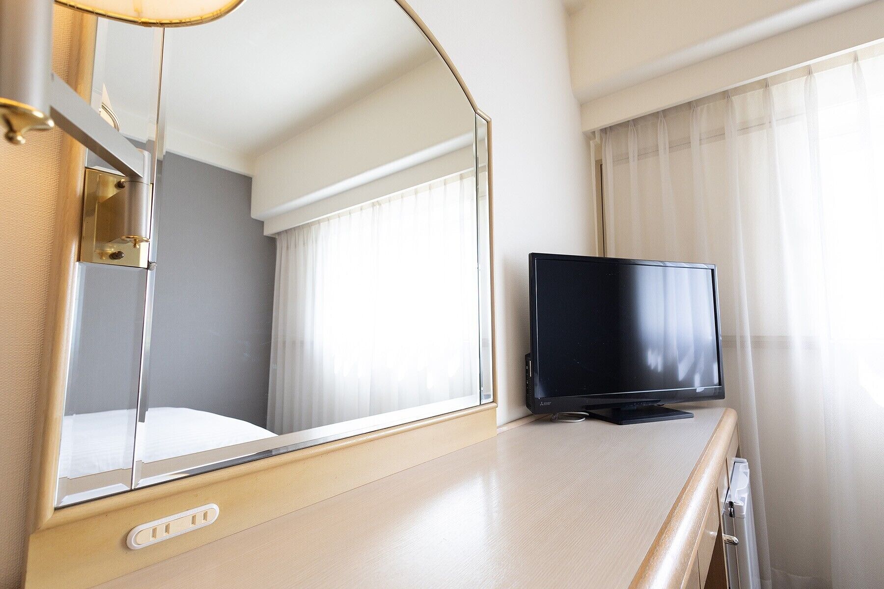 A hotel room with a bed, a television on a wooden dresser, a mirror, and a window with curtains.