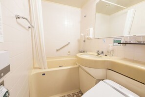 Une petite salle de bain avec une baignoire, un lavabo et des toilettes. Il y a une barre d’appui dans la zone de la douche et un miroir au-dessus du lavabo.