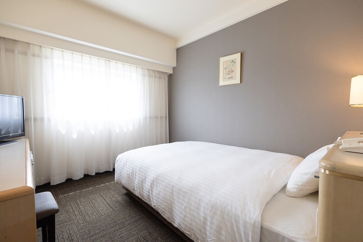 Une chambre d’hôtel comprenant un lit, un bureau avec une télévision, une table de chevet avec une lampe et une fenêtre avec des rideaux légers.