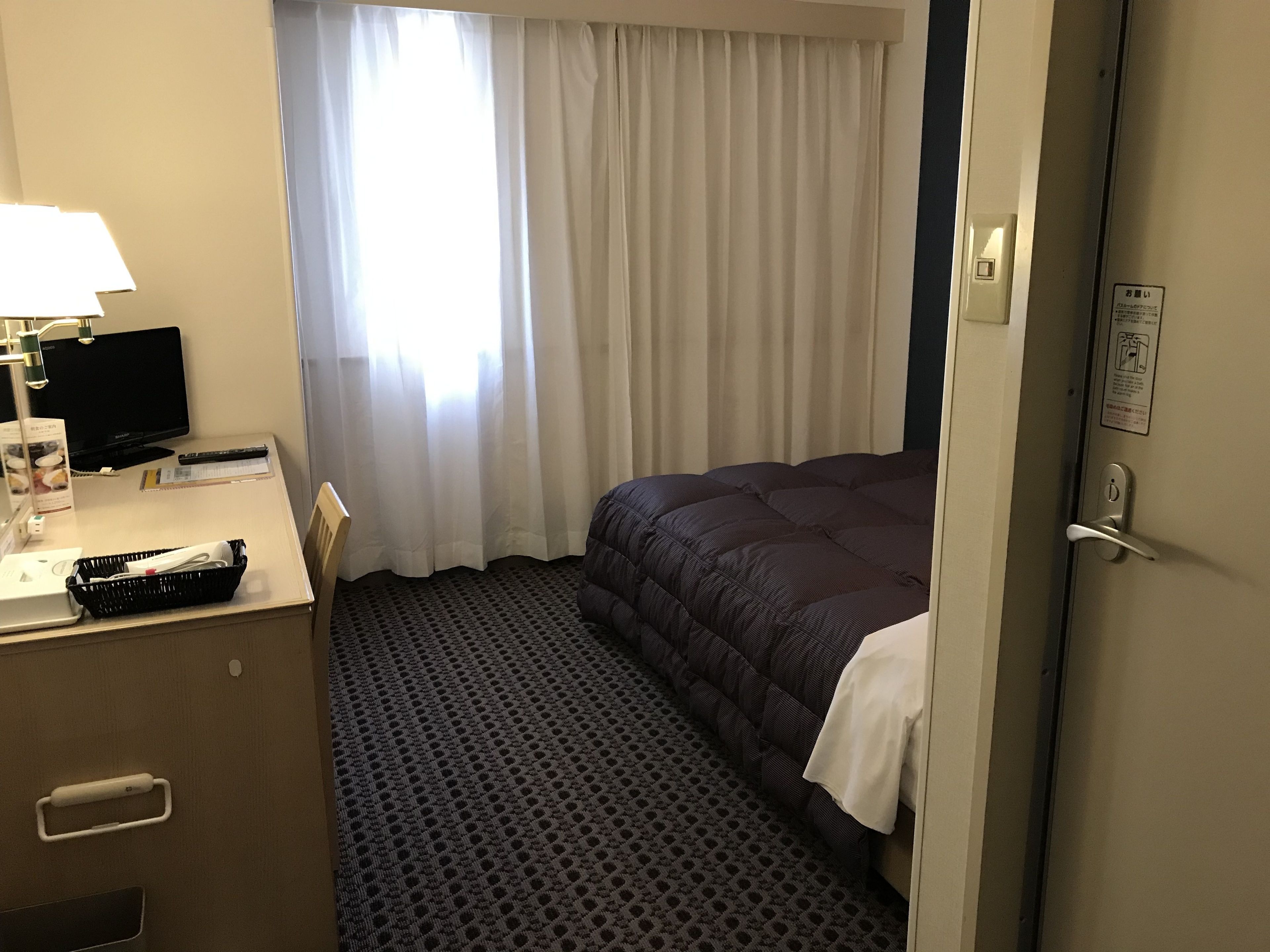 A hotel room with a bed, a desk with a lamp, a television, and a window with curtains.