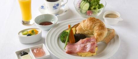 Un desayuno con croissant, jamón, huevos en un plato, vaso de zumo de naranja, café, ensalada y recipientes pequeños de mermelada y mantequilla.