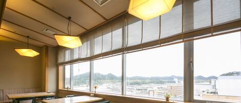Un comedor con mesas y sillas, ventanas amplias con vistas a la ciudad y montañas, y lámparas colgantes.