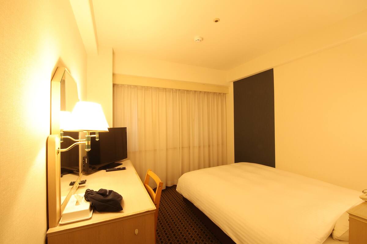 Hotelkamer met een bed, een bureau met lamp, een stoel, een televisie en een raam met gordijnen.