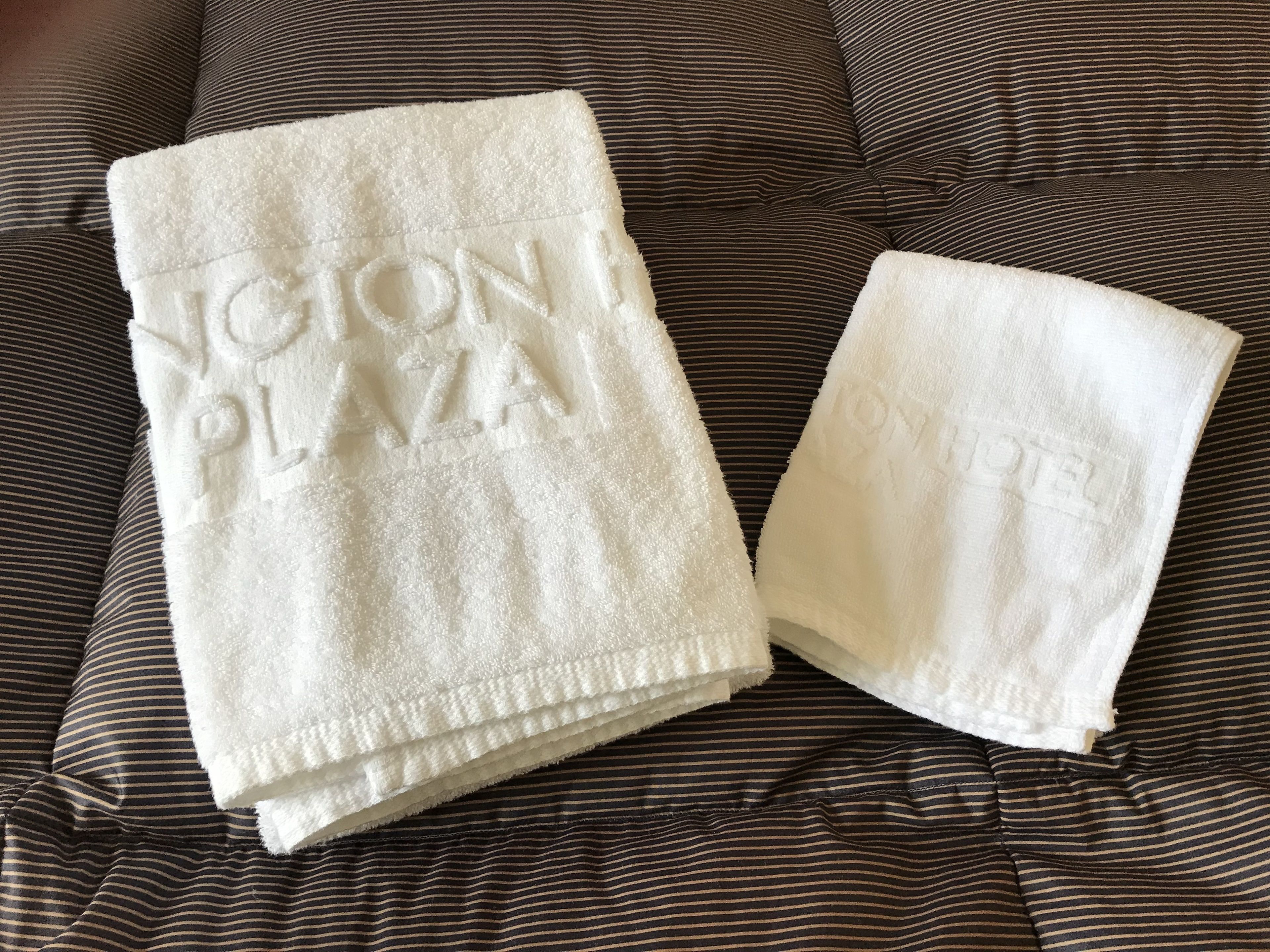 Two white towels with "Región Plaza" embossed on them, placed on a striped bed.