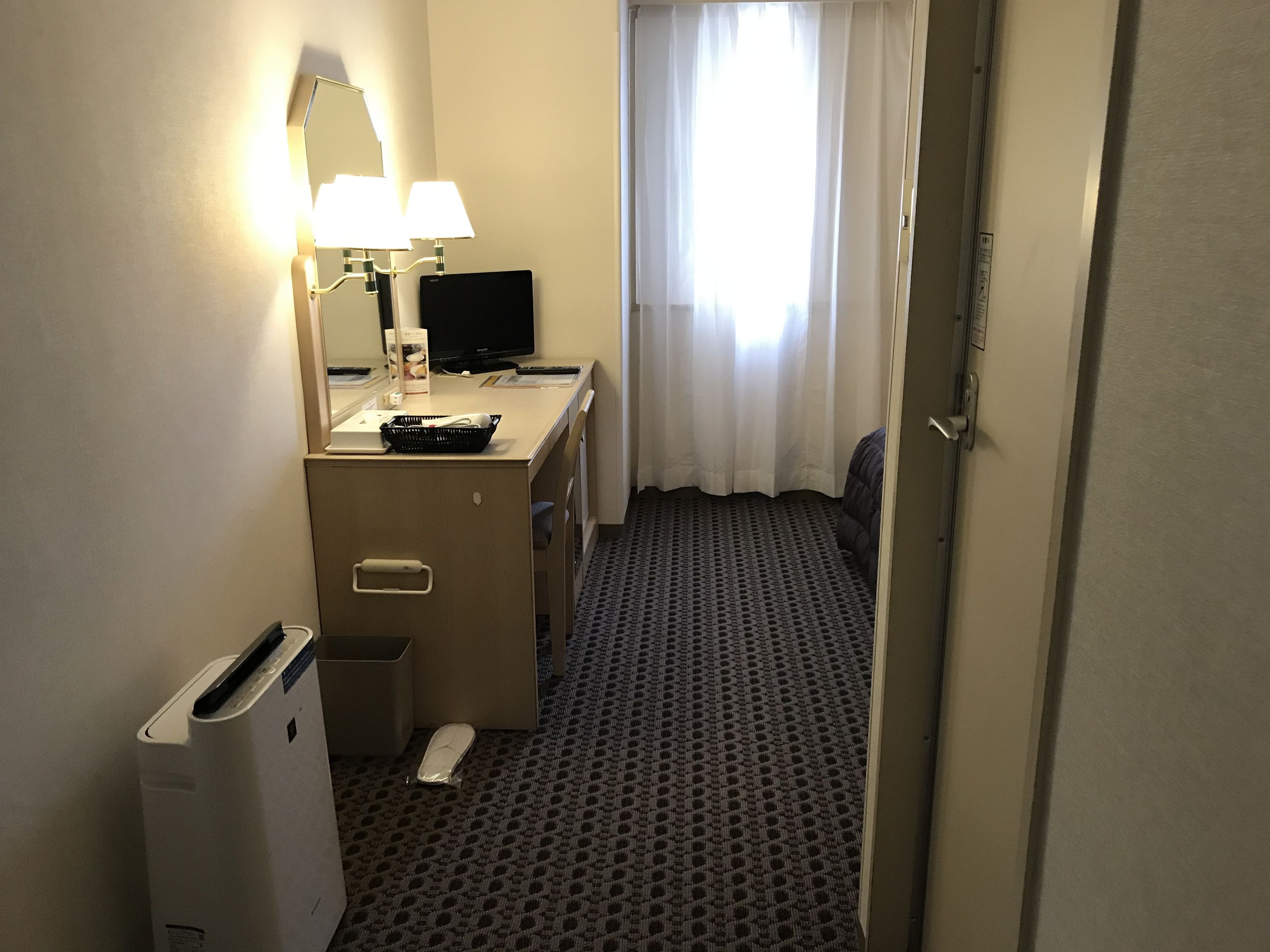 A hotel room with a desk, a computer, a lamp, a bed, and a trash bin.