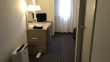 A hotel room with a desk, a computer, a lamp, a bed, and a trash bin.