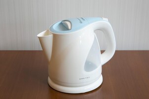 Una tetera eléctrica blanca con tapa y asa azules, sobre una mesa de madera frente a una pared blanca.