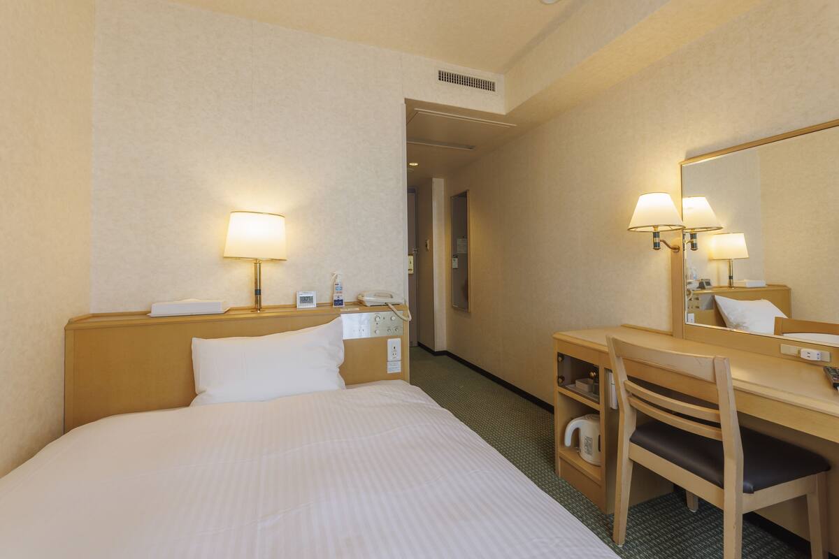 A hotel room with a bed, bedside table, lamp, chair, desk, and mirror.