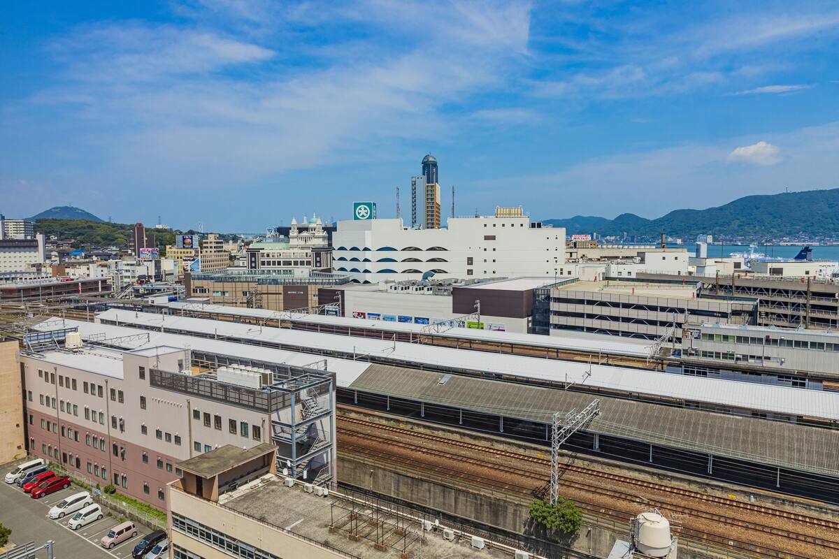 A train station with multiple tracks and platforms, surrounded by industrial buildings and a mountainous backdrop.