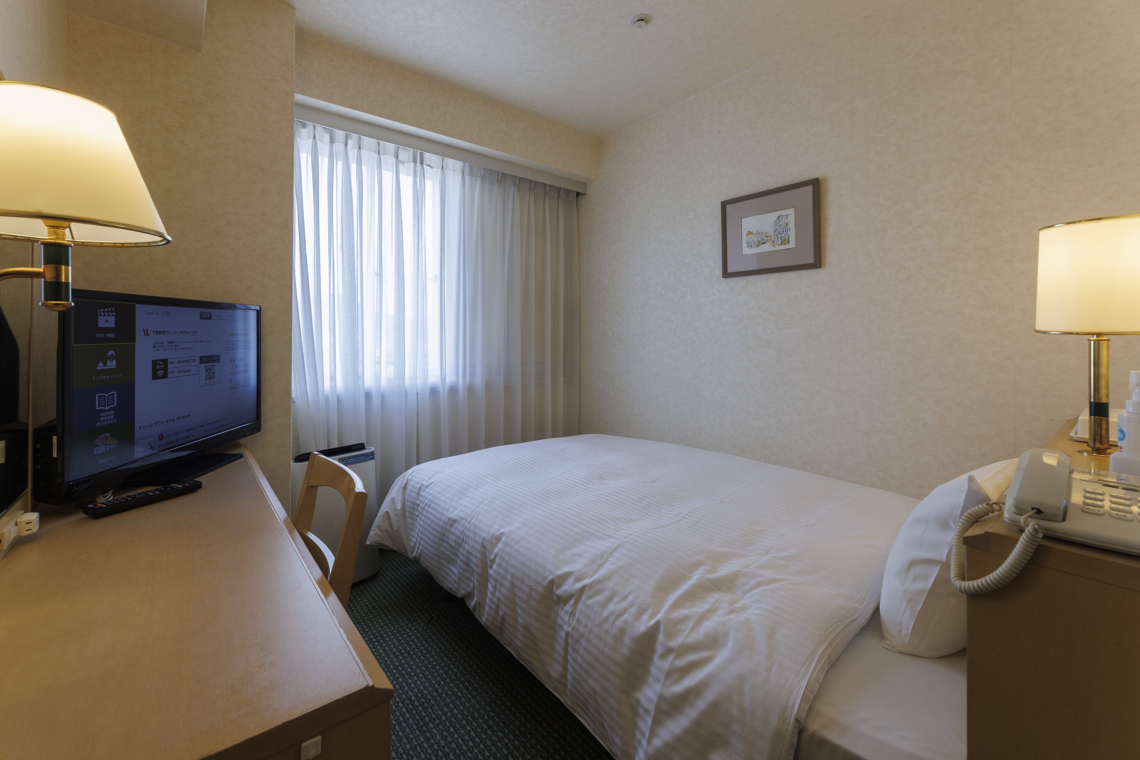 Habitación de hotel con una cama, un escritorio con computadora y lámpara, un teléfono y una ventana con cortinas.