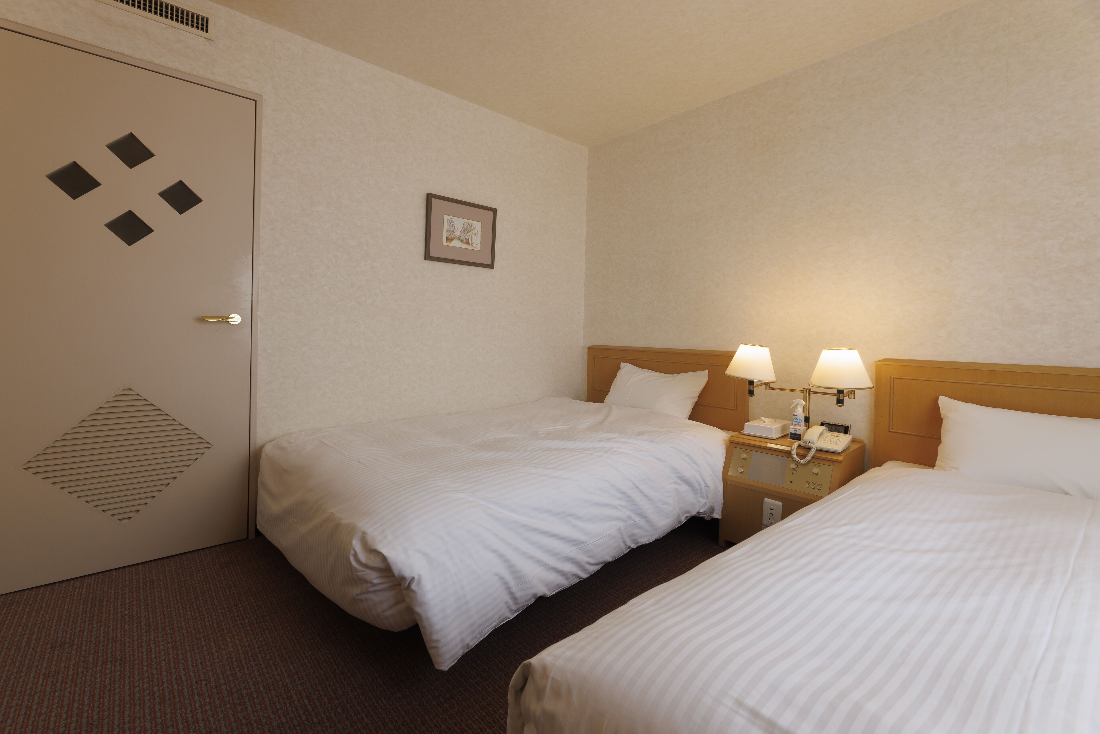 Habitación de hotel con dos camas, una puerta con diseño geométrico, un cuadro enmarcado en la pared y una mesita de noche con lámparas y un teléfono.
