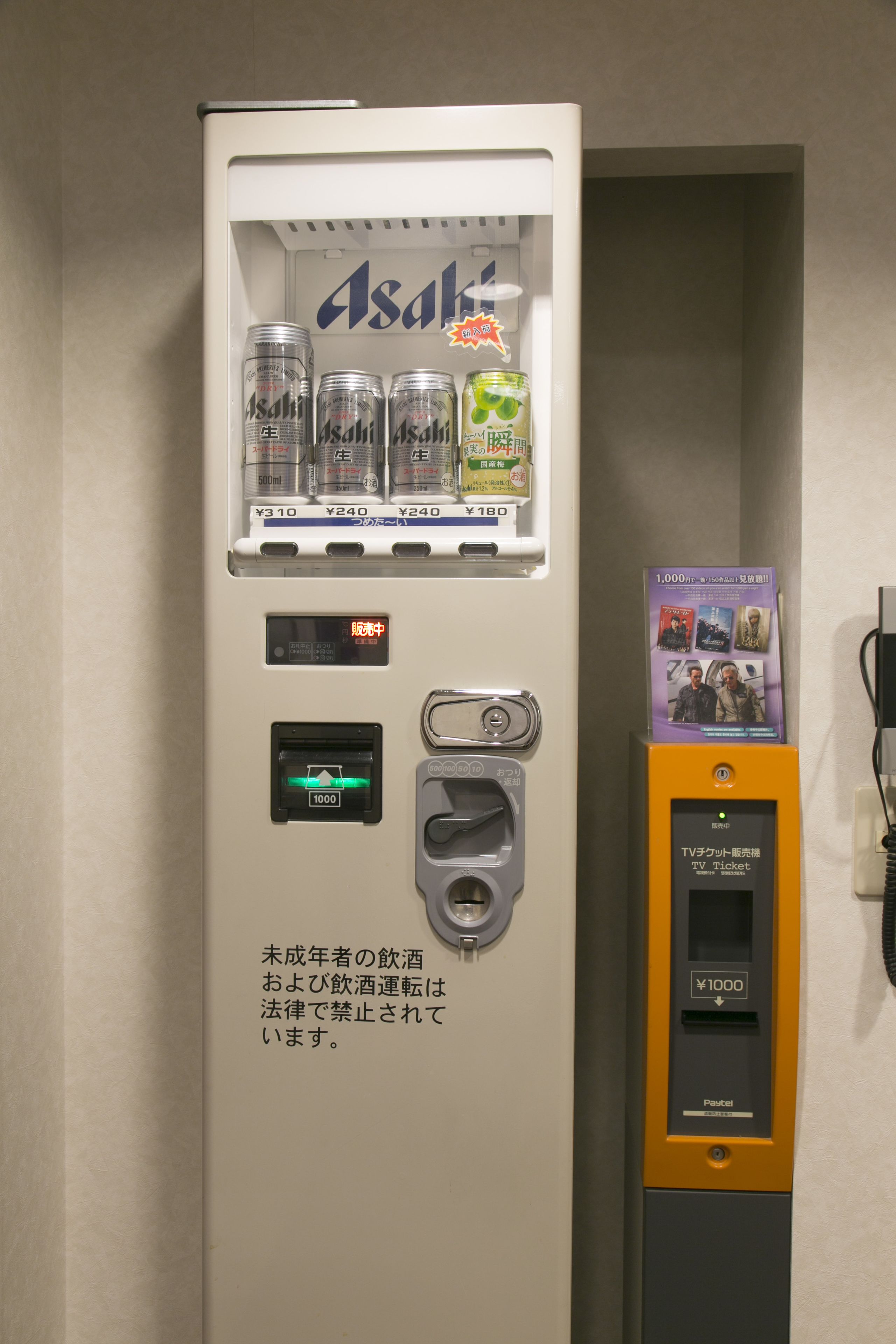 Una macchina distributrice con lattine di birra Asahi, una macchina per l'acquisto di biglietti televisivi e un cartello con testo in giapponese.
