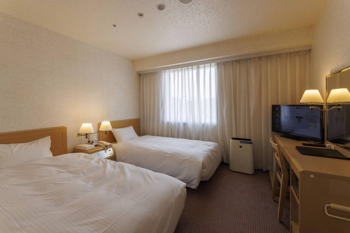 A hotel room with two beds, a desk with a computer, a TV, and a window with curtains.