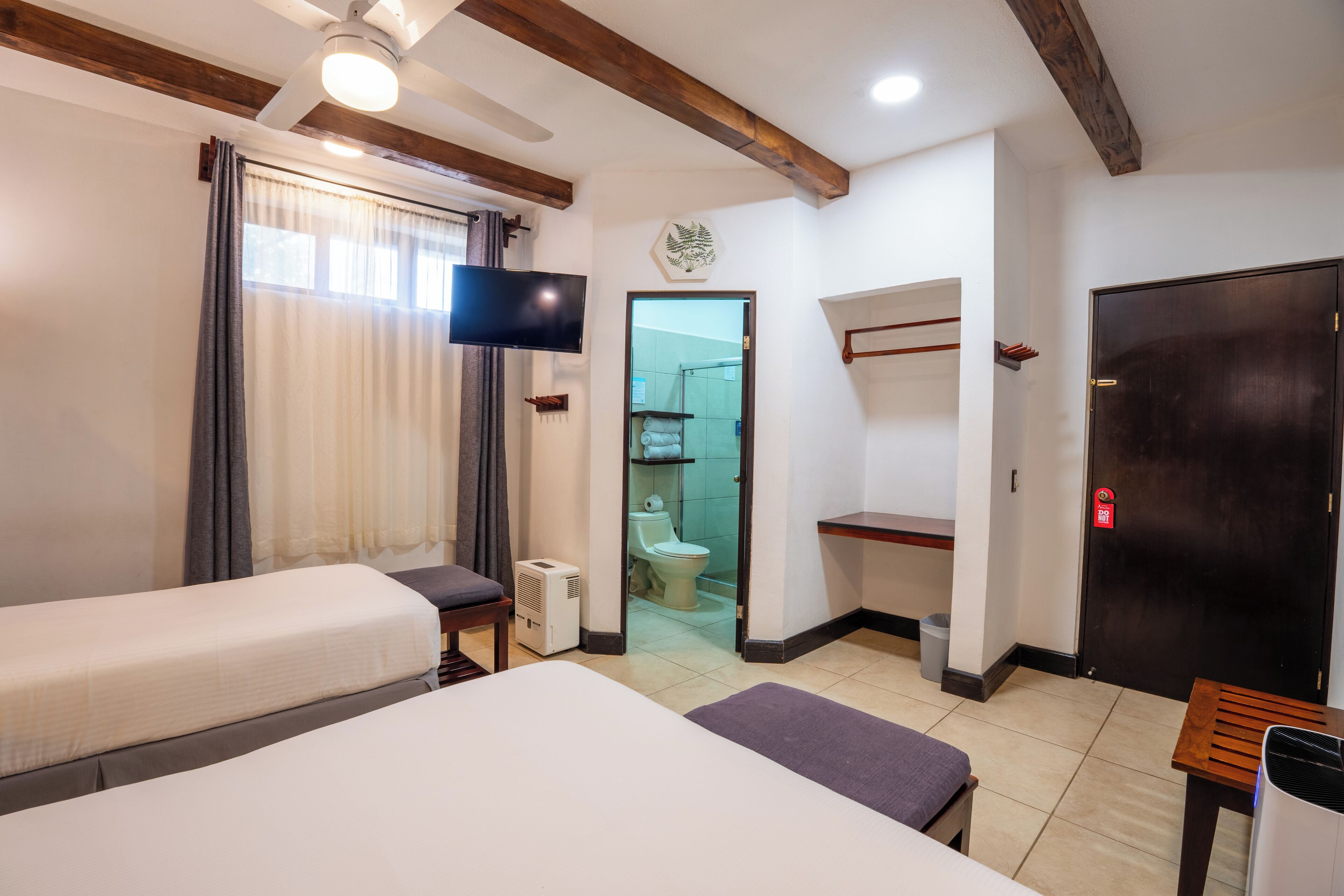Une chambre d’hôtel avec deux lits, une télévision, un ventilateur de plafond, un plafond avec poutres apparentes, une salle de bain avec des toilettes et une douche, et une porte avec un panneau rouge indiquant la sortie de secours.