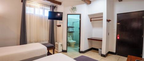 A hotel room with two beds, a television, a ceiling fan, a wooden beam ceiling, a bathroom with a toilet and shower, and a door with a red emergency exit sign.