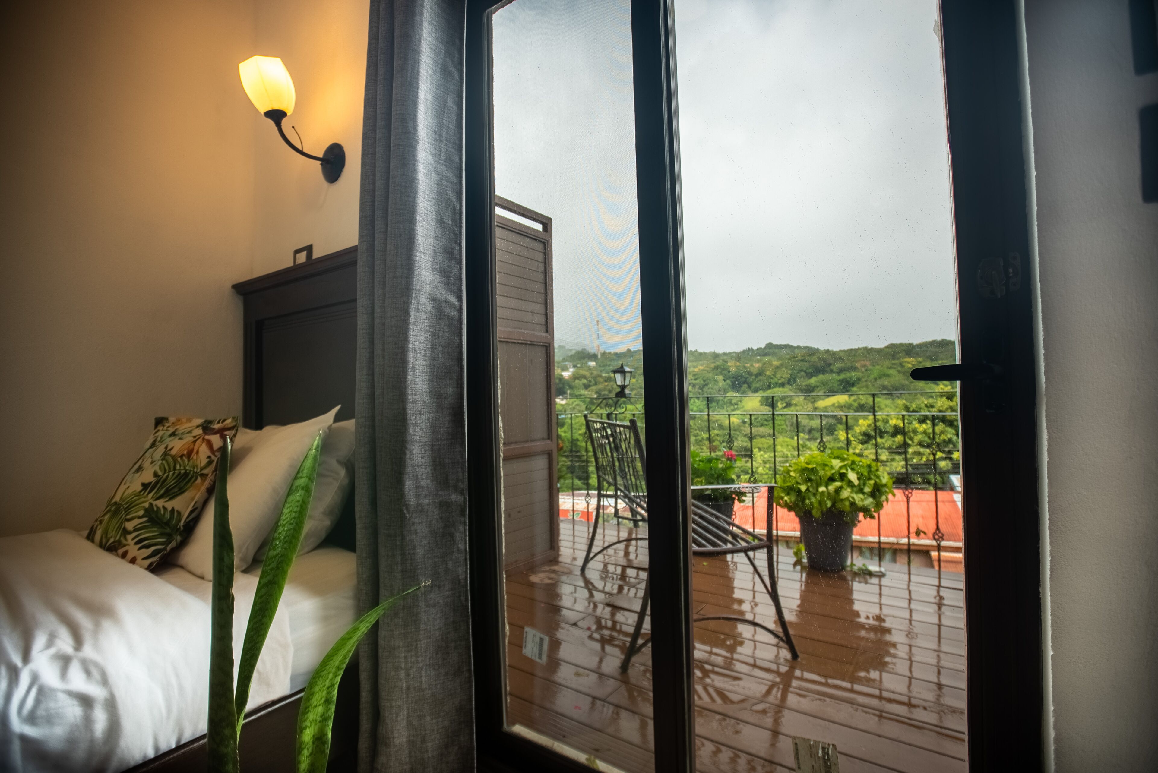 Een slaapkamer met een bed, een nachtkastje en een raam met uitzicht op een balkon met een stoel en een potplant.
