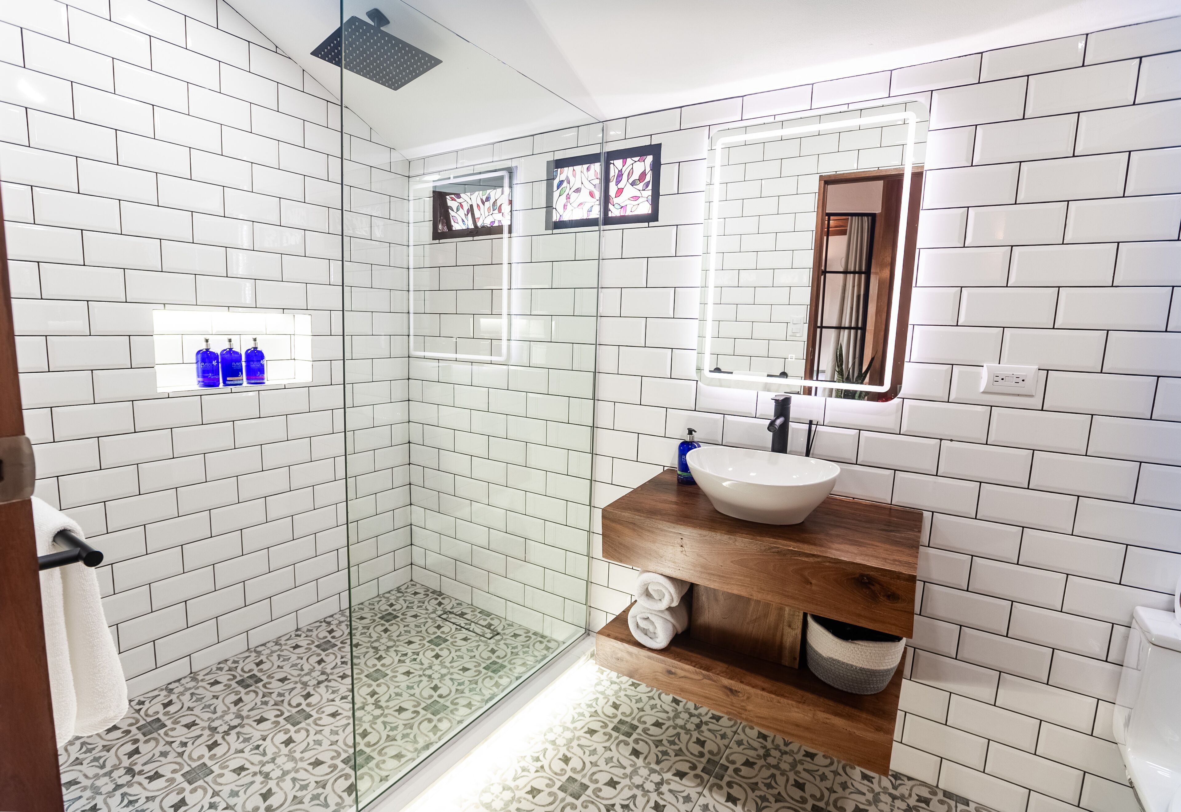 A modern bathroom with white subway tiles, a glass shower enclosure, a wooden vanity with a sink, and a mirror with stained glass.