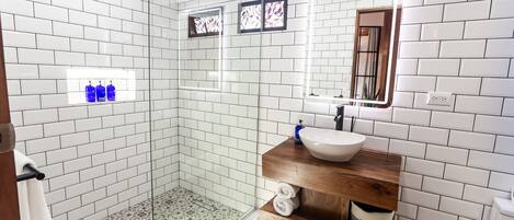 A modern bathroom with white subway tiles, a glass shower enclosure, a wooden vanity with a sink, and a mirror with stained glass.