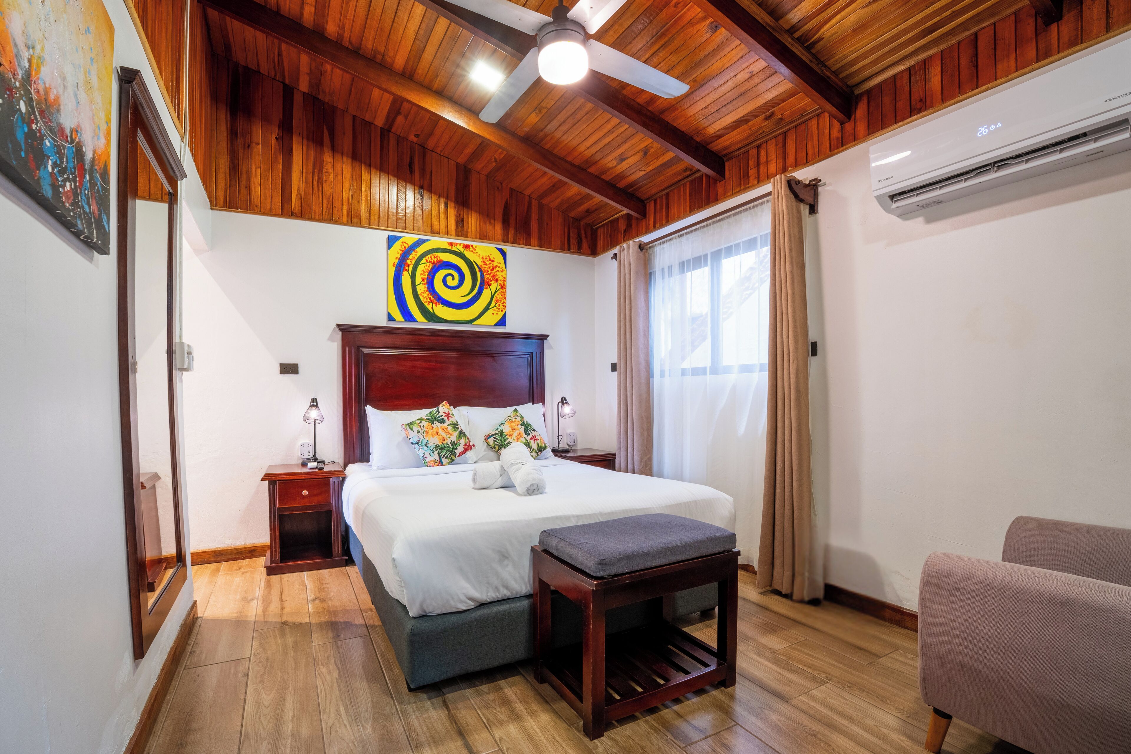 A hotel room with a wooden ceiling, a bed with a headboard, a bedside table with a lamp, a small wooden stool, and a chair.