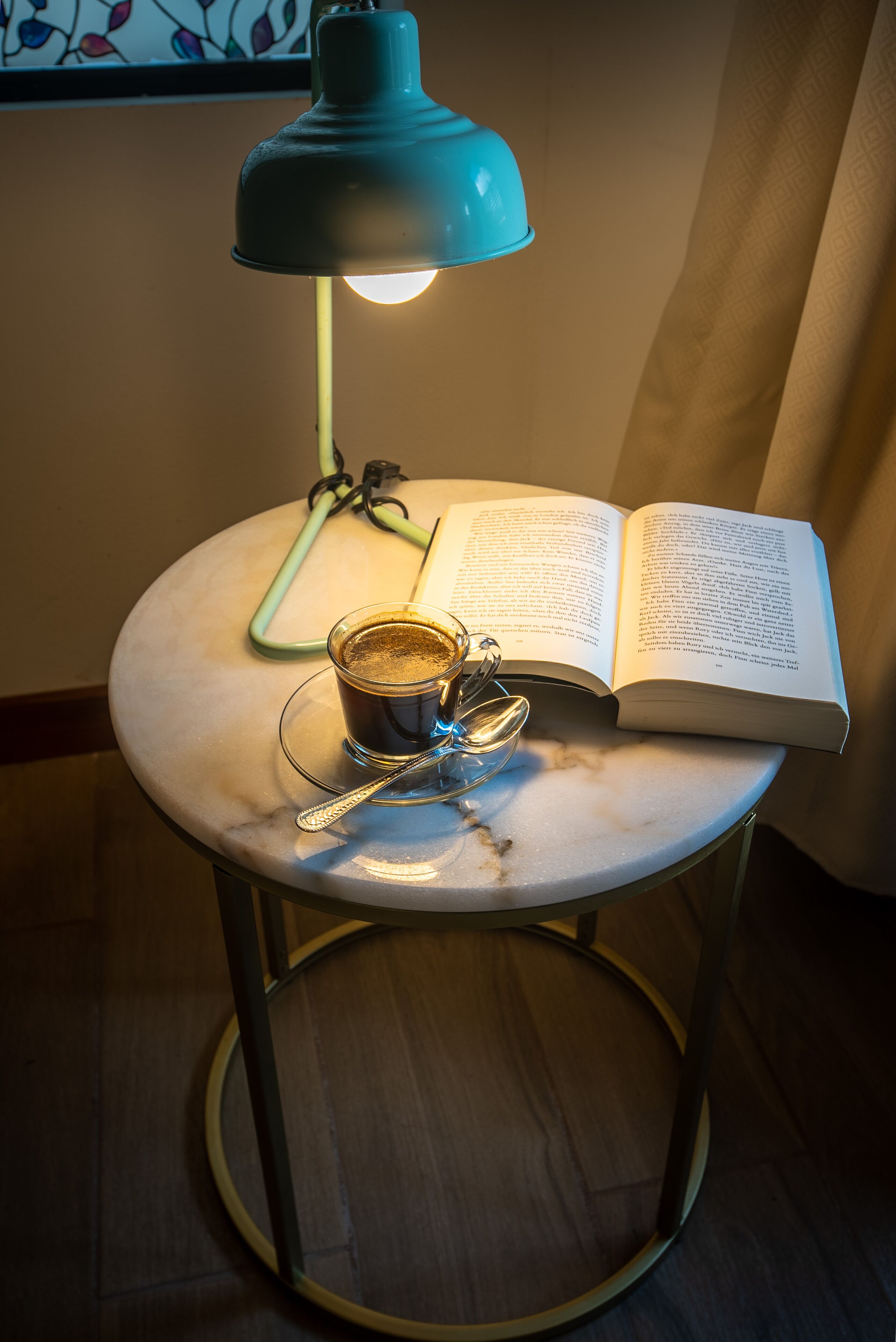 A round table with a book, a cup of coffee, and a spoon. A blue lamp is on the table, and a green pen is on the book.