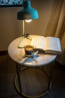 Una mesa redonda con un libro, una taza de café y una cuchara. Una lámpara azul está sobre la mesa, y una pluma verde sobre el libro.