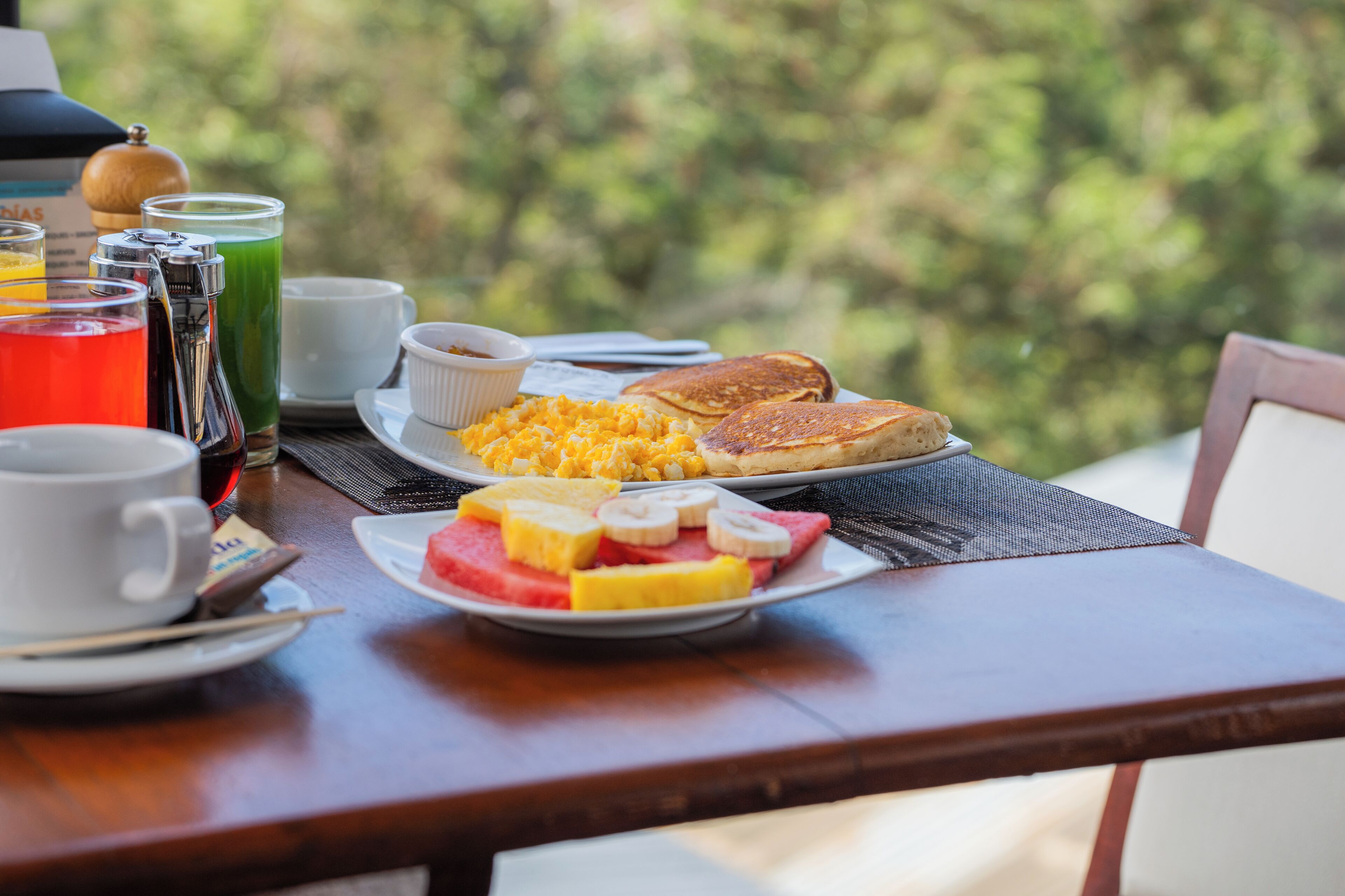 A breakfast table with plates of eggs, pancakes, and fruit, and a variety of beverages.