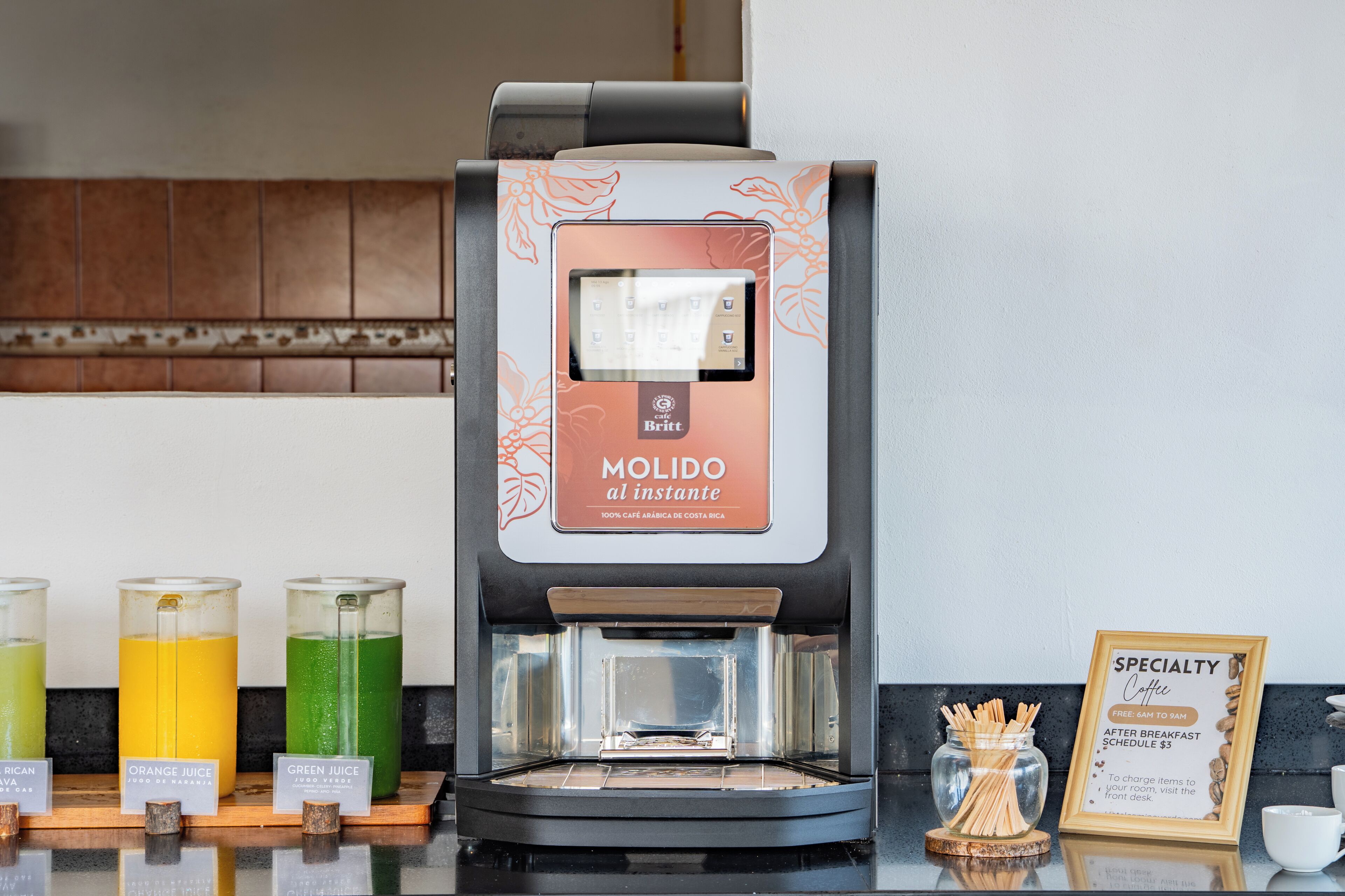 A coffee machine with a digital interface, three labeled juice dispensers, and a framed sign about specialty coffee.