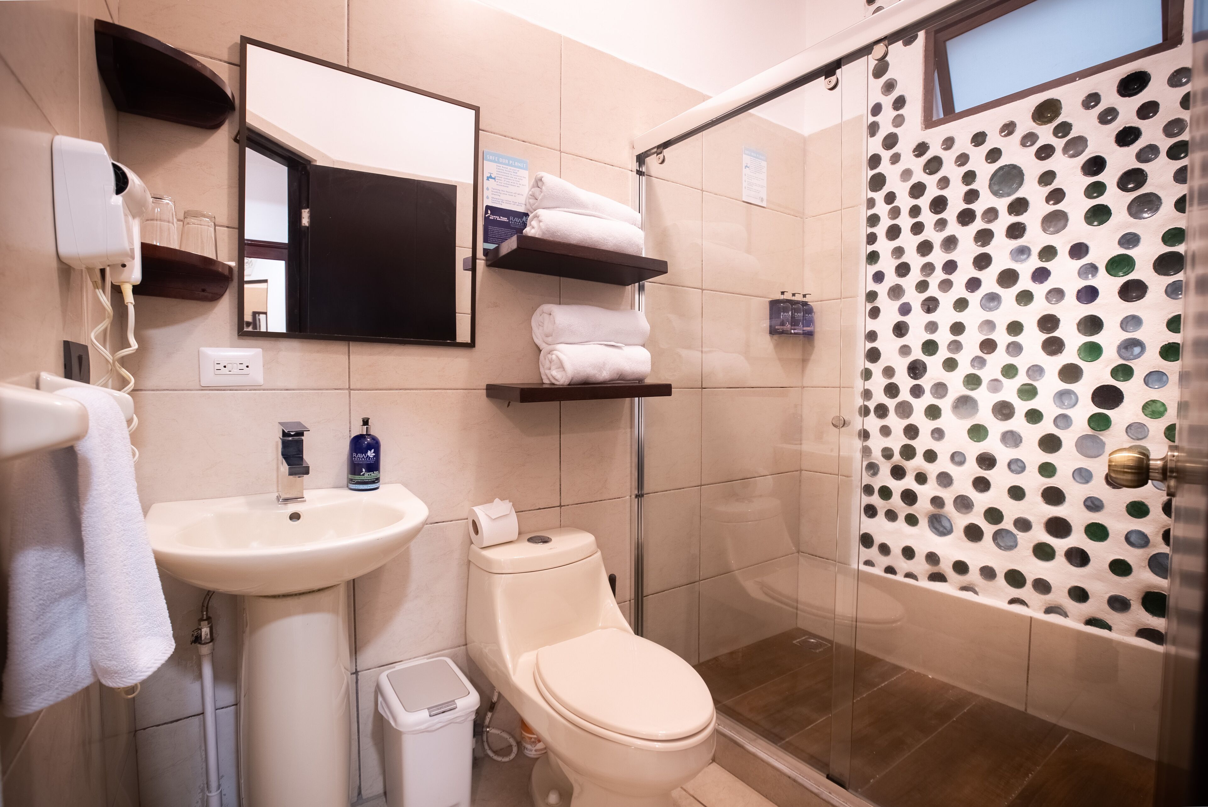 Une salle de bain avec un lavabo, des toilettes et une douche. On y trouve des serviettes sur une étagère, un sèche-cheveux et un miroir.