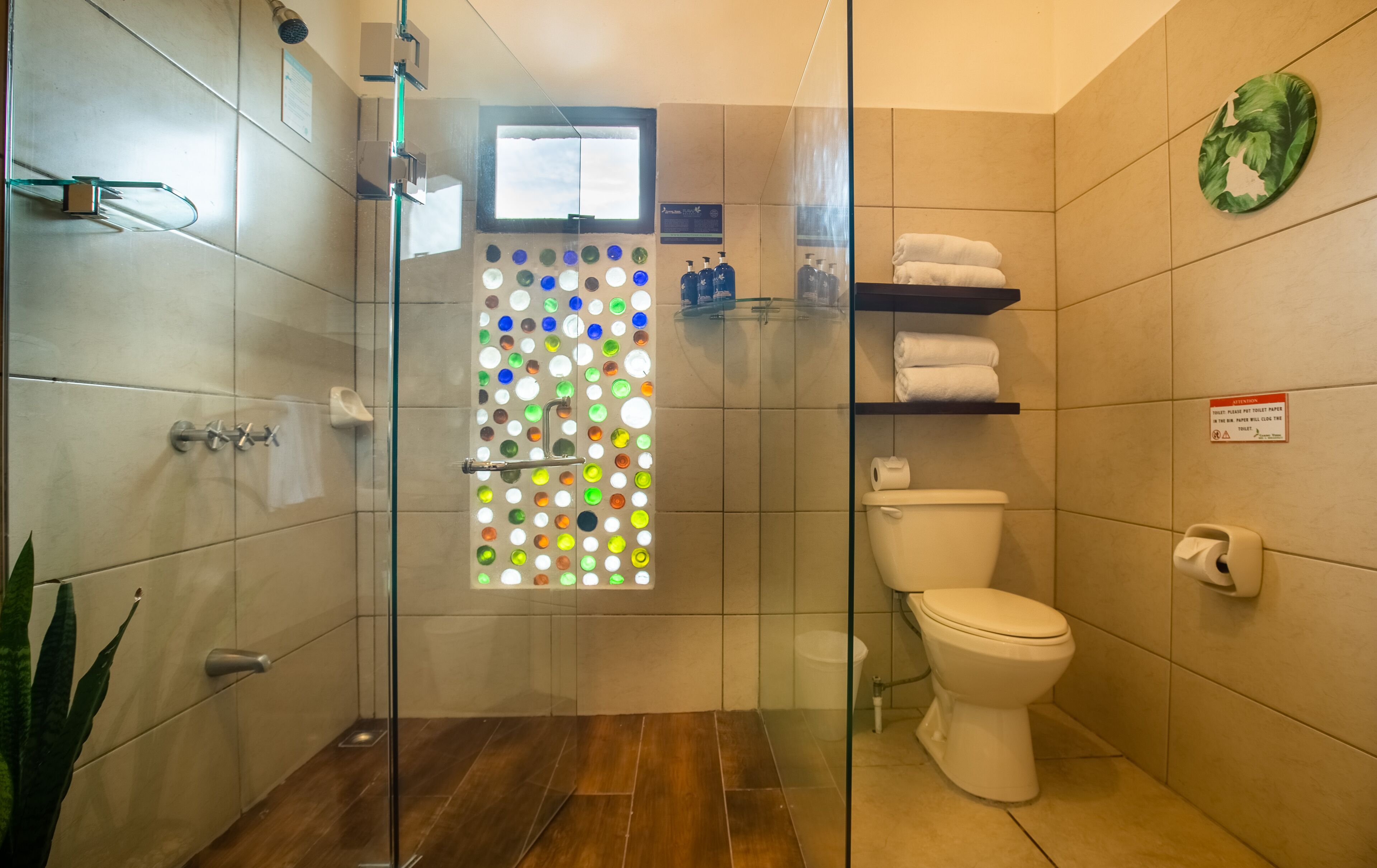 Ein Badezimmer mit Duschkabine aus Glas, Toilette, Ablagefläche mit Handtüchern und einer dekorativen Glasscheibe mit farbigen Kreisen.