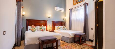 A hotel room with two beds, a small table, a bench, a patterned rug, and a painting on the wall.