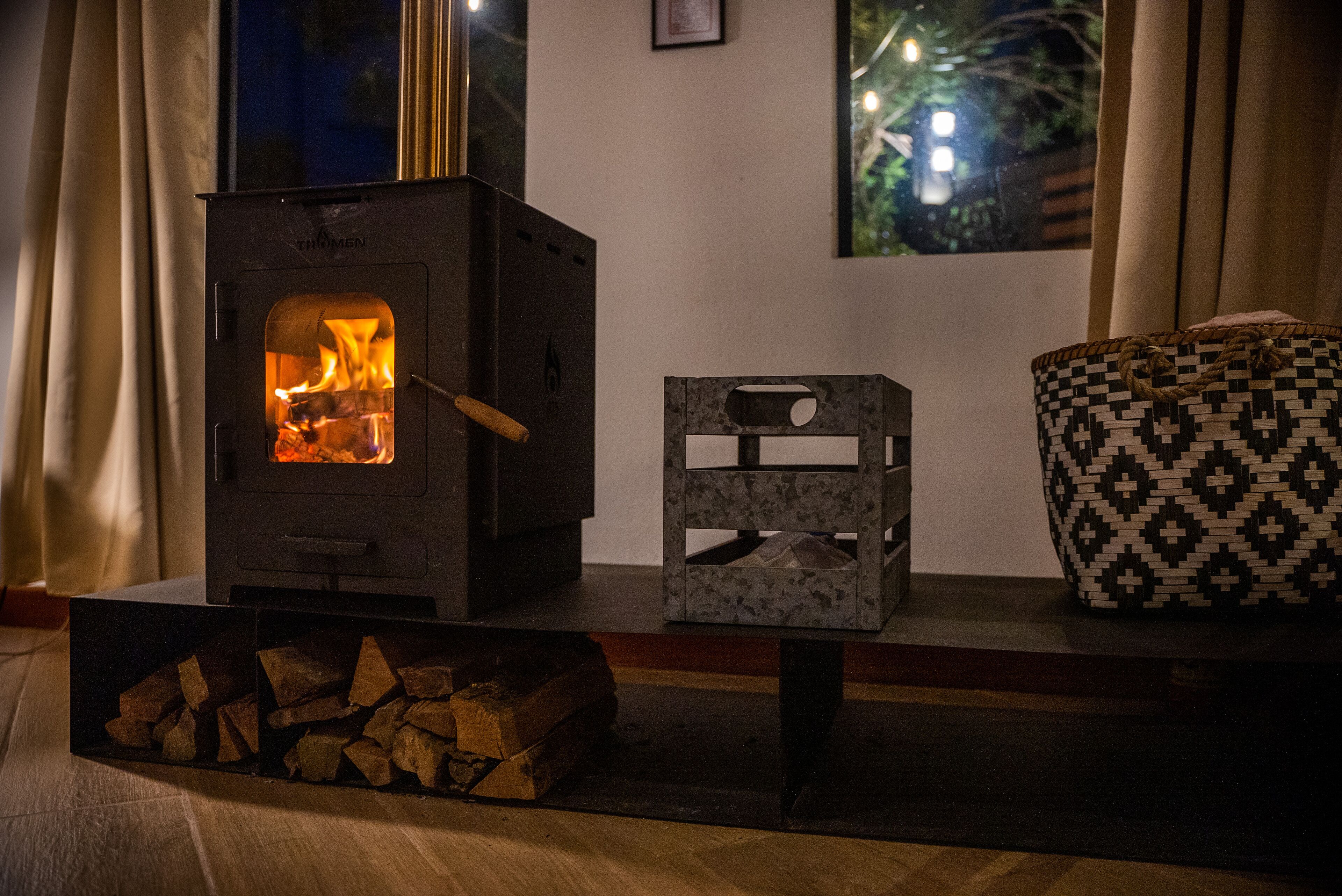 A wood-burning stove with visible flames, a patterned basket, and a stack of firewood.