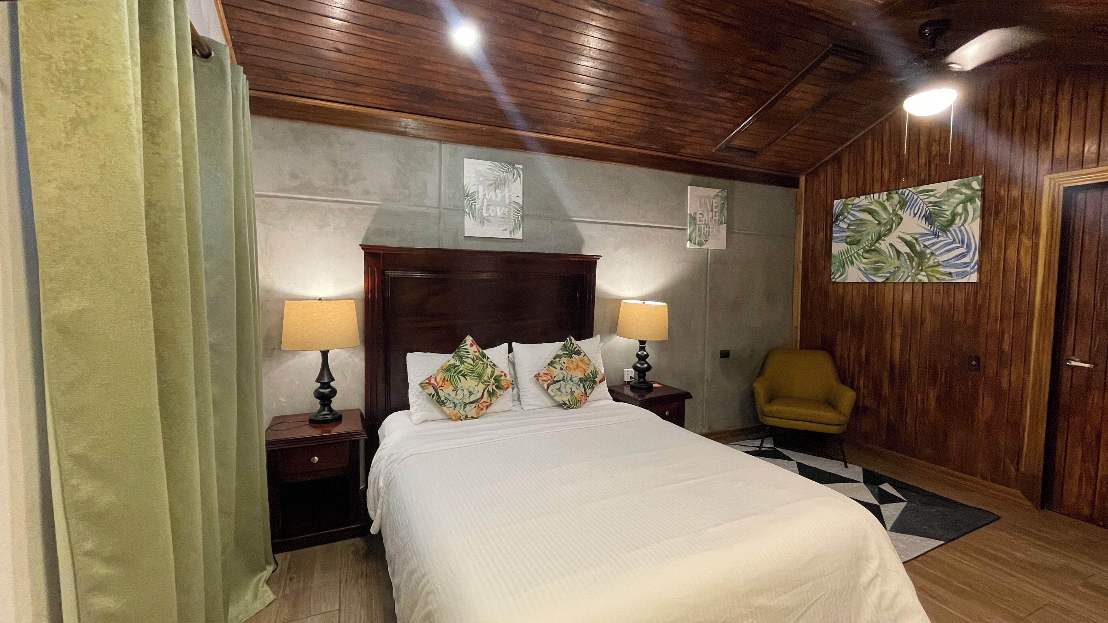 Un dormitorio con cabecera de madera, dos lámparas de noche, un cojín estampado con flores, un sillón amarillo y un cuadro en la pared.