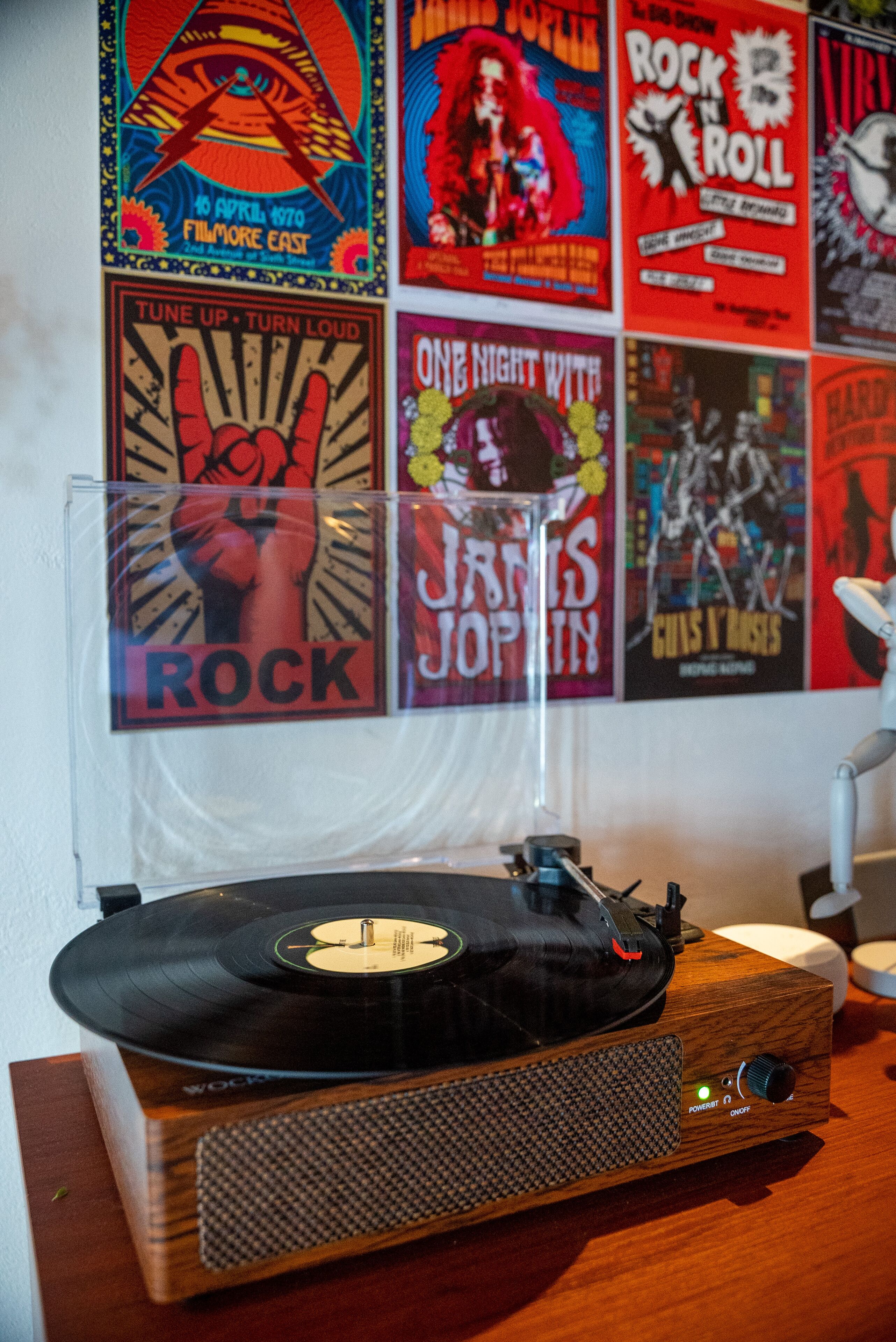 Een houten draaitafel met een vinylplaat, een bureaulamp en een muur met verschillende posters van rockbands.