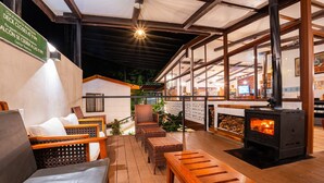 A cozy outdoor seating area with wicker furniture, a wood-burning stove, and a view of the interior through large windows.