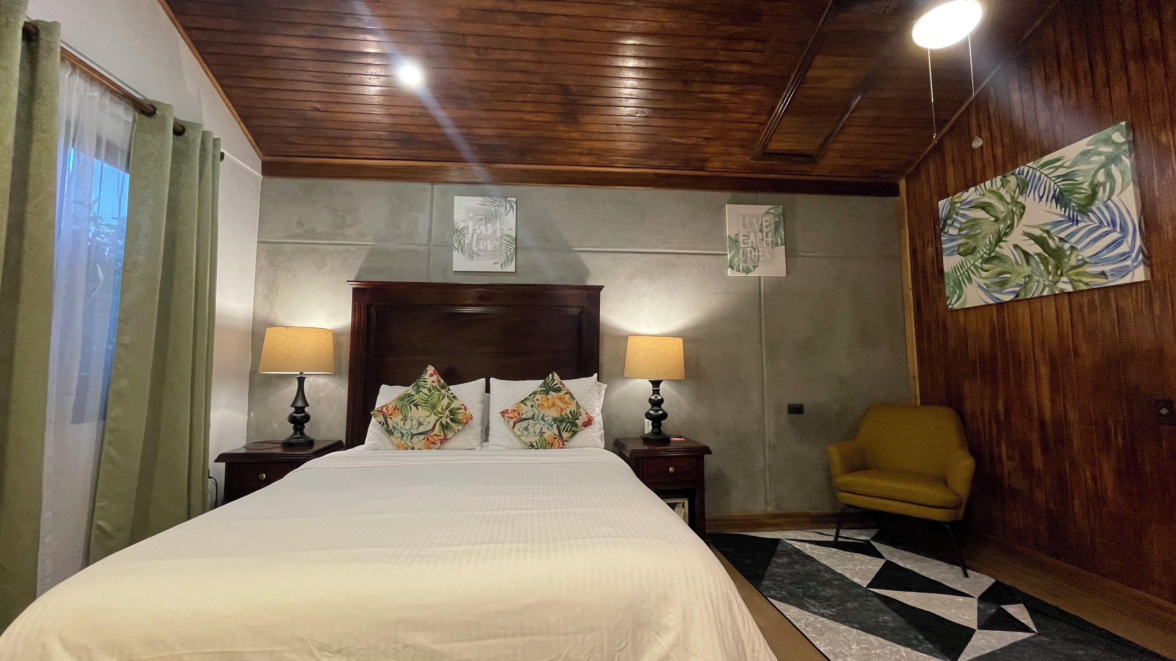 Ein Schlafzimmer mit einem großen Bett, zwei Nachttischlampen, einem Holz-Kopfteil, einem gelben Sessel und einem Wandbild mit tropischem Motiv.